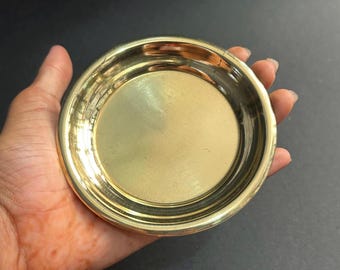 May include: A small, round, brass-coloured dish with a slightly raised rim. The dish has a smooth, reflective surface and is held in a hand against a dark background. The dish appears to be made of metal.