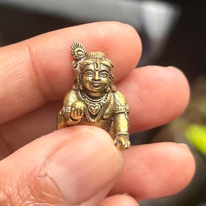 May include: A small, gold-colored metal figurine of a Hindu deity, Krishna, with a feather headdress and a heart-shaped pendant. The figurine is sitting with its legs crossed and its hands clasped in front of it.