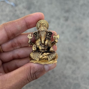 Peut inclure: Une petite statue en métal dorée de la divinité hindoue Ganesha. La statue représente Ganesha assis, sa trompe levée, tenant un trident et une hache. La statue est finement détaillée et possède une auréole autour de la tête.