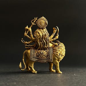 May include: A gold-colored statue of a deity riding a lion. The deity has multiple arms and wears a detailed headdress. The lion is adorned with an ornate saddle. The statue is set against a dark background.