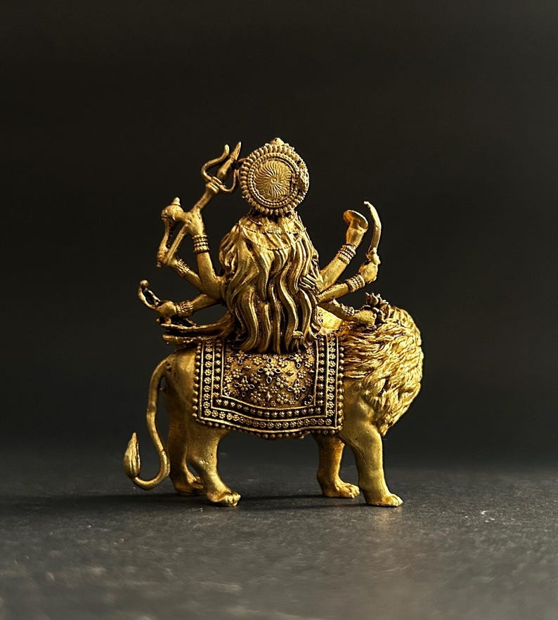 May include: A gold-colored statue of a deity riding a lion. The deity has multiple arms and wears a detailed headdress. The lion is adorned with an ornate saddle. The statue is set against a dark background.