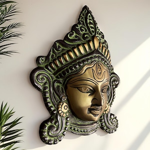 May include: A bronze-coloured wall hanging of a deity's face. The face has a serene expression, with detailed features and a third eye. The surrounding frame is dark green with gold accents, and the piece is mounted on a white wall.