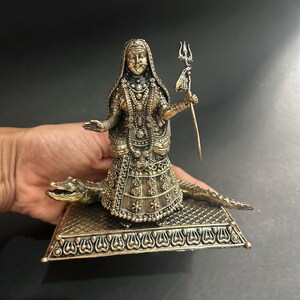 K&ouml;nnte beinhalten: Eine detaillierte Bronzestatue einer Gottheit, geschm&uuml;ckt mit aufwendigem Schmuck und Kopfschmuck. Die Figur steht auf einem Krokodil und h&auml;lt einen Dreizack. Die Basis der Statue hat ein verziertes Design.