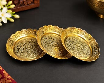May include: Three circular gold-colored metal plates featuring an intricate embossed floral pattern across the base and a scalloped edge. The plates have a polished finish and are arranged in a slightly overlapping row on a dark surface.
