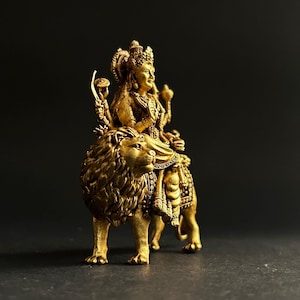 May include: A golden statue of a deity riding a lion. The statue is intricately detailed, with the deity wearing a crown and ornate jewelry. The lion is also highly detailed, with a flowing mane and a regal pose. The statue is set against a dark background.