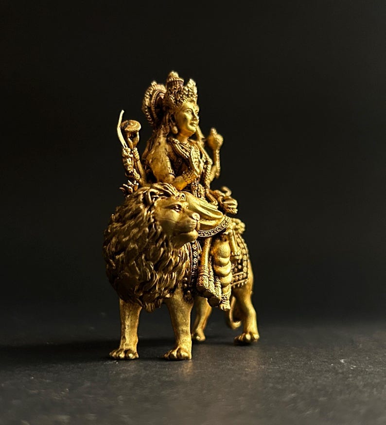 May include: A golden statue of a deity riding a lion. The statue is intricately detailed, with the deity wearing a crown and ornate jewelry. The lion is also highly detailed, with a flowing mane and a regal pose. The statue is set against a dark background.