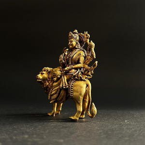 May include: A golden statue of a deity riding a lion. The figure is adorned with intricate details, including a crown and multiple arms. The lion is also intricately detailed, with a flowing mane and a powerful stance. The statue is set against a dark background.