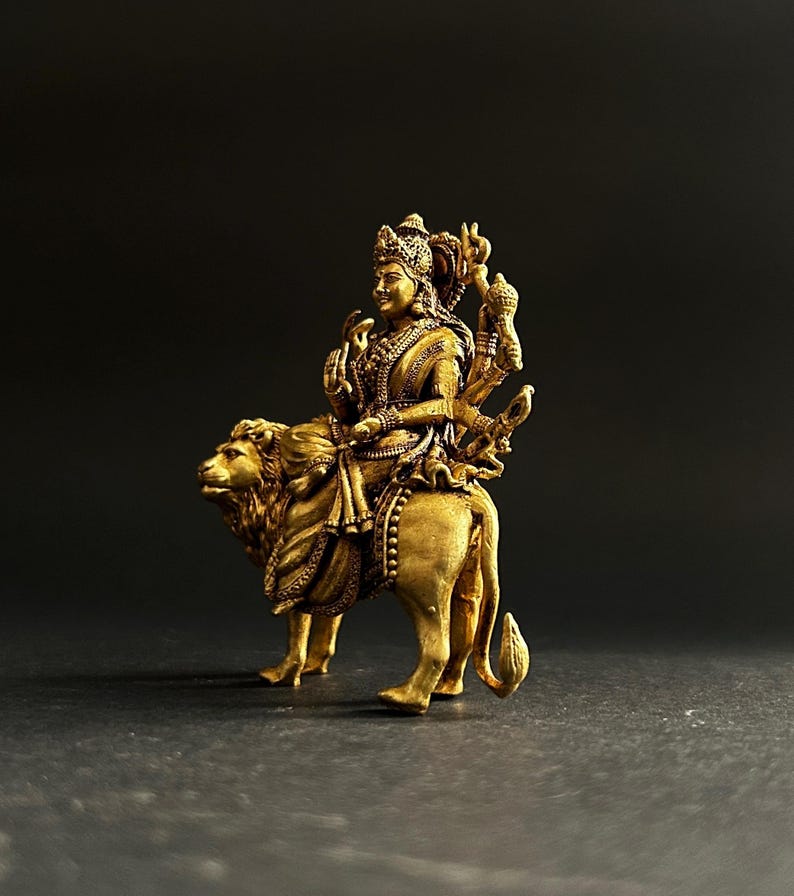 May include: A golden statue of a deity riding a lion. The figure is adorned with intricate details, including a crown and multiple arms. The lion is also intricately detailed, with a flowing mane and a powerful stance. The statue is set against a dark background.