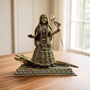 K&ouml;nnte beinhalten: Eine detaillierte Bronzestatue einer Gottheit, geschm&uuml;ckt mit aufwendigem Schmuck und Kopfschmuck, die einen Dreizack h&auml;lt. Die Figur steht auf einem Krokodil, das auf einem dekorativen Sockel montiert ist. Die Statue steht auf einem Holztisch.