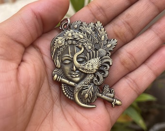 May include: A gold-toned metal pendant featuring a detailed depiction of a Hindu deity with a peacock feather headdress and a flute. The pendant is intricately designed with a peacock feather motif and a crescent moon.