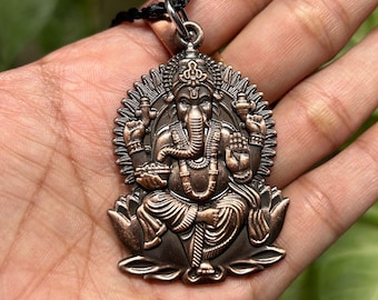 May include: A copper-toned pendant necklace featuring a detailed depiction of the Hindu deity Ganesha. The pendant shows Ganesha sitting on a lotus flower with his trunk raised and holding a sweet in his hand. The pendant is surrounded by a decorative halo.