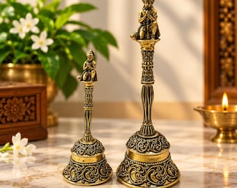 May include: Two antique-style brass bells with ornate floral engravings on the base. Each bell features a handle topped with a small, detailed figurine of a kneeling deity with hands pressed together in a prayer position.