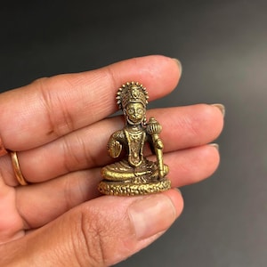 Puede incluir: Una pequeña estatua de metal dorada de una deidad hindú sentada. La estatua está detallada con intrincados tallados y presenta una corona y un bastón.