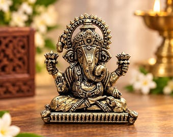 May include: A metallic gold-toned statue of the deity Ganesha in a seated position. The figure features an elephant head, multiple arms holding lotus flowers, and an ornate crown. It rests on a rectangular base with detailed carvings.