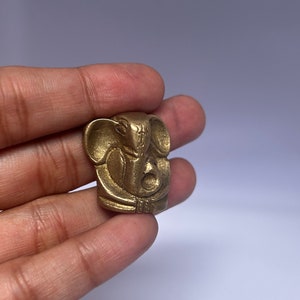 Puede incluir: Una pequeña estatua de bronce dorada de la deidad hindú Ganesha. La estatua es detallada y muestra a Ganesha sentado con la trompa levantada.