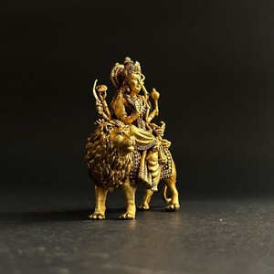 May include: A gold-colored statue of a deity riding a lion. The deity is adorned with intricate details, including a crown and multiple arms holding various objects. The lion has a detailed mane and is in a walking pose. The statue is set against a dark background.