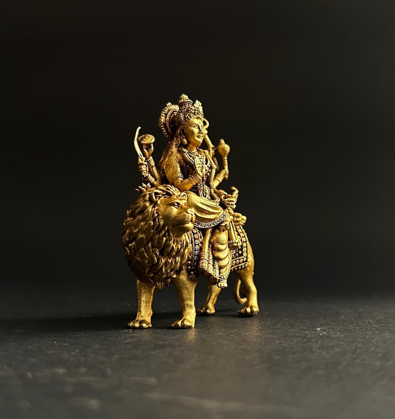 May include: A gold-colored statue of a deity riding a lion. The deity is adorned with intricate details, including a crown and multiple arms holding various objects. The lion has a detailed mane and is in a walking pose. The statue is set against a dark background.