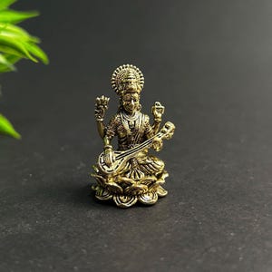 Op de afbeelding: Een klein, gedetailleerd messing beeld van een zittende godheid die een snaarinstrument bespeelt. De figuur is versierd met ingewikkelde sieraden en een halo, zittend op een lotusbloem basis. Goudkleurig, donkere achtergrond.