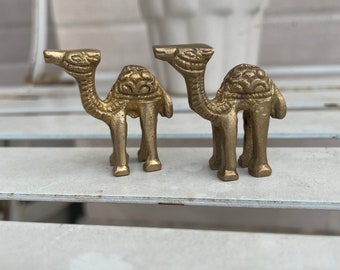 May include: Two gold-colored metal camel figurines. The camels are standing on a white surface.