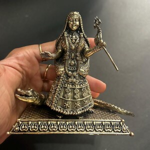 K&ouml;nnte beinhalten: Eine detaillierte Statue in Bronzeoptik einer Gottheit, geschm&uuml;ckt mit aufwendigem Schmuck und einem drapierten Gewand. Die Figur h&auml;lt einen Dreizack und steht auf einer Plattform mit Krokodilbasis. Die Statue wird von einer Hand gehalten.