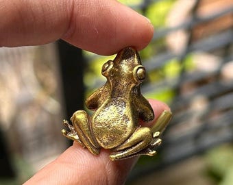 Pode incluir: Uma pequena figura de sapo de cor dourada com uma superfície texturizada. O sapo está sentado sobre suas patas traseiras com as patas dianteiras estendidas.