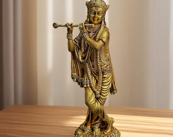May include: A gold-coloured statue of a deity playing a flute. The figure is adorned with detailed jewellery and a crown, standing on a textured base. The statue is set on a wooden surface, with a neutral background.