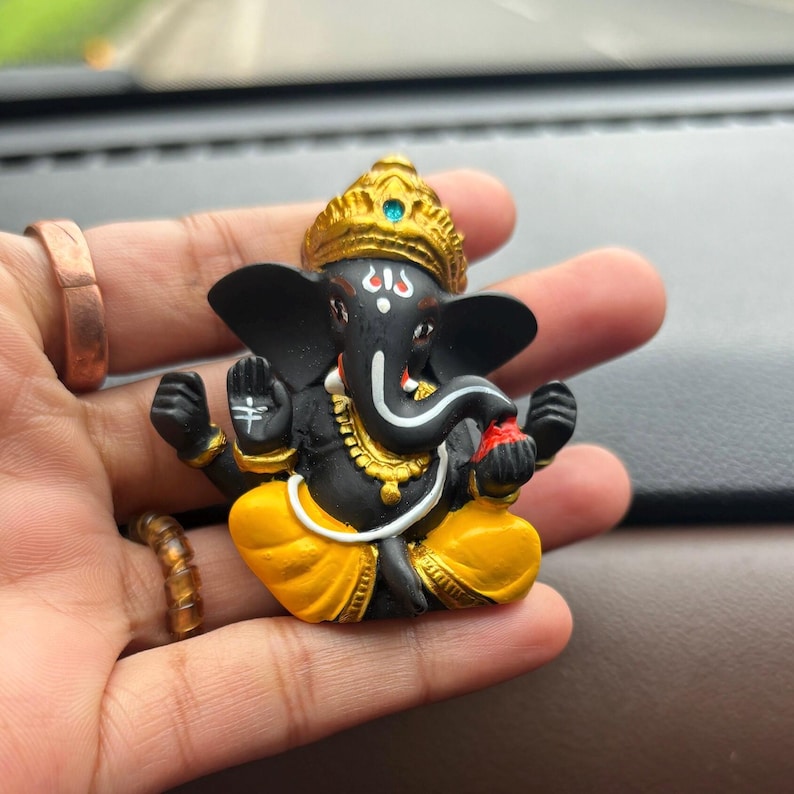 Puede incluir: Una peque&ntilde;a figura negra de la deidad hind&uacute; Ganesha. La figura lleva un dhoti amarillo y dorado y una corona dorada. La figura sostiene un peque&ntilde;o objeto rojo en su mano derecha.