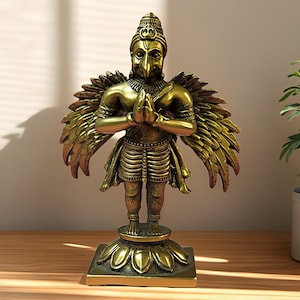 May include: A gold-coloured statue of a deity with a bird-like head and human body, hands clasped in prayer. The statue has detailed wings and is mounted on a lotus flower base. The statue is displayed on a wooden surface.