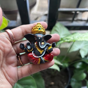 Puede incluir: Figura de Ganesha negra con una corona dorada y un dhoti rojo y dorado. La figura sostiene un objeto peque&ntilde;o en su mano derecha y tiene una trompa blanca.