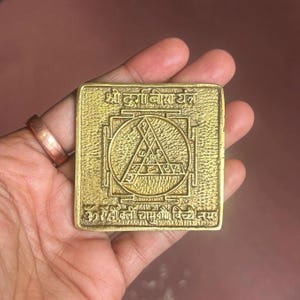 Puede incluir: Una placa cuadrada de metal dorado con una inscripción en sánscrito y un diseño geométrico. La inscripción dice "Shri Durga Visarjan" y "Om Hreem Kleem Chamundaye Vichche Namaha".
