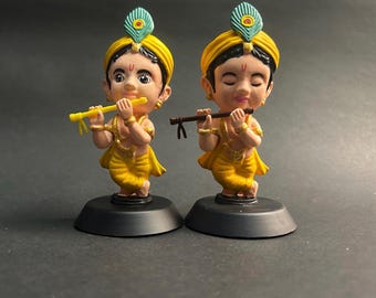 May include: Two figurines of a young person in yellow clothing, playing a flute. Each figure has a yellow turban with a peacock feather and stands on a black base. The background is a dark gray.
