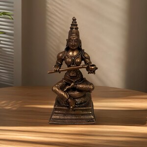 May include: A bronze-colored statue of a seated figure playing a flute. The figure is adorned with detailed clothing and jewelry, and sits on a detailed base. The statue is displayed on a wooden surface, with a neutral background.
