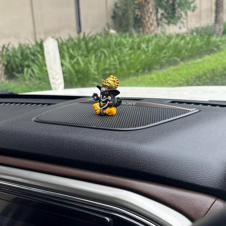 Puede incluir: Una peque&ntilde;a figura negra y dorada del dios hind&uacute; Ganesha, sentado en una postura meditativa. La figura est&aacute; colocada en el tablero de un coche, delante de un altavoz negro con la etiqueta "SONY".