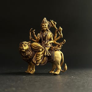 May include: A detailed golden statue of a deity riding a lion. The deity has multiple arms and is adorned with intricate jewelry and a crown. The lion is also highly detailed, with a textured mane and body. The statue is set against a dark background.