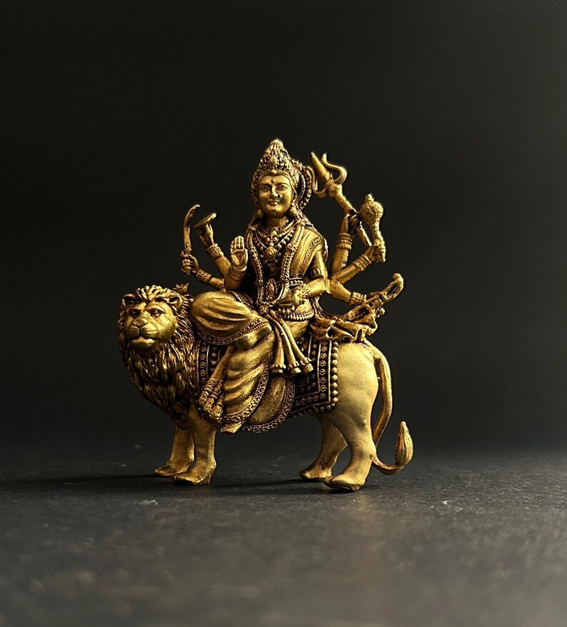 May include: A detailed golden statue of a deity riding a lion. The deity has multiple arms and is adorned with intricate jewelry and a crown. The lion is also highly detailed, with a textured mane and body. The statue is set against a dark background.
