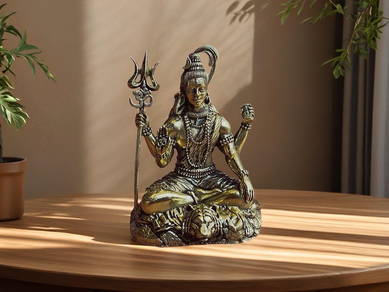 Pode incluir: Uma est&aacute;tua de metal em tom dourado da divindade Shiva sentada em posi&ccedil;&atilde;o meditativa sobre uma pele de tigre. A figura segura um tridente com um s&iacute;mbolo Om, possui m&uacute;ltiplos bra&ccedil;os, usa colares de contas e uma lua crescente no cabelo.