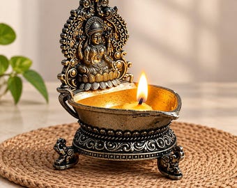 May include: A metallic oil lamp featuring an ornate backplate with a relief of a seated figure on a lotus base. The bowl has a gold-toned interior and holds a lit tea light candle. The base is supported by three decorative feet.