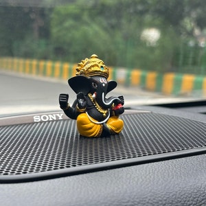 Puede incluir: Una peque&ntilde;a figura negra y dorada del dios hind&uacute; Ganesha, sentada en el tablero de instrumentos de un coche negro con una rejilla de altavoz negra. La figura lleva una t&uacute;nica amarilla y una corona dorada.