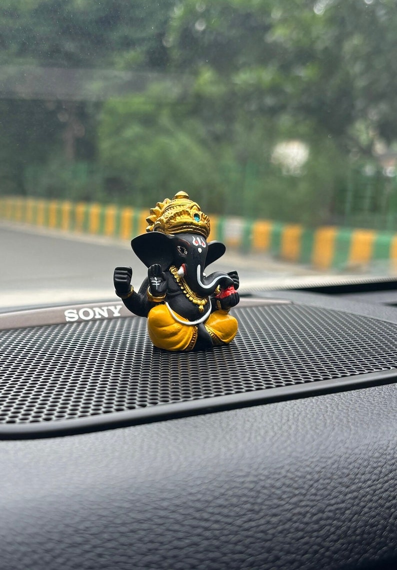 Puede incluir: Una peque&ntilde;a figura negra y dorada del dios hind&uacute; Ganesha, sentada en el tablero de instrumentos de un coche negro con una rejilla de altavoz negra. La figura lleva una t&uacute;nica amarilla y una corona dorada.