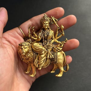 May include: A gold-colored statue of a deity seated on a lion. The deity has multiple arms and is adorned with intricate jewelry and a crown. The lion is detailed with a flowing mane and a saddle. The statue is held in a hand against a dark background.