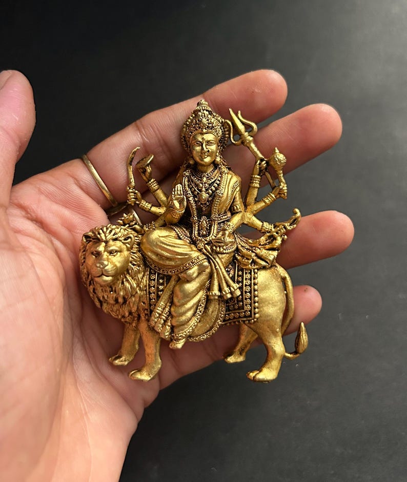 May include: A gold-colored statue of a deity seated on a lion. The deity has multiple arms and is adorned with intricate jewelry and a crown. The lion is detailed with a flowing mane and a saddle. The statue is held in a hand against a dark background.