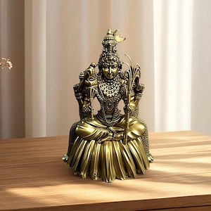 Puede incluir: Una estatua dorada detallada de una deidad sentada, adornada con intrincadas joyas y un tocado alto. La figura sostiene objetos en sus manos y está sentada sobre una base decorativa. La estatua está colocada sobre una superficie de madera.