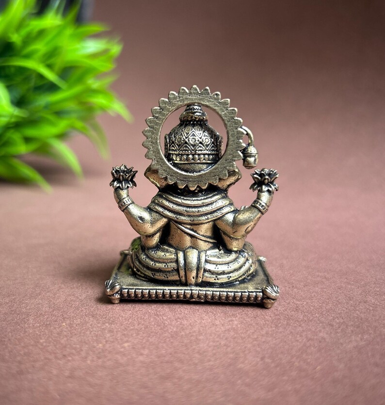 May include: A gold-colored metal statue of Ganesha, the Hindu god of wisdom and prosperity, sitting on a decorative base. The statue is detailed with intricate carvings and features a halo behind the head.