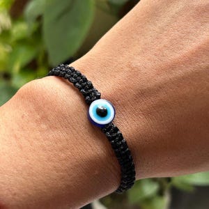 Pode incluir: Uma pulseira de corda preta com um amuleto de olho turco azul. O amuleto tem um centro branco e uma pupila preta.