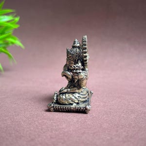 May include: Silver-toned metal figurine of Ganesha, the Hindu god of wisdom and prosperity, sitting on a square base. The figurine is intricately detailed with a crown, a trunk, and a flower in its hand.