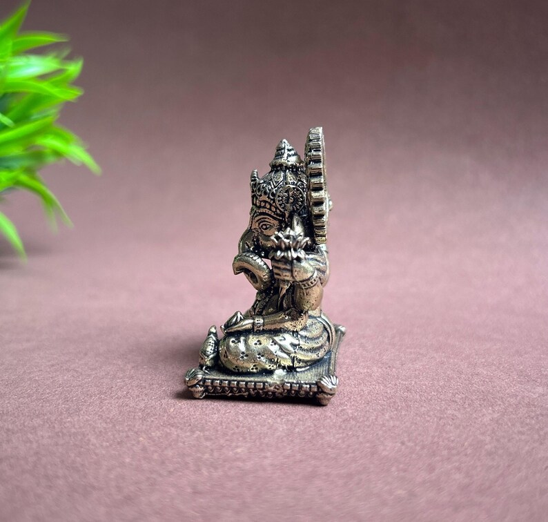May include: Silver-toned metal figurine of Ganesha, the Hindu god of wisdom and prosperity, sitting on a square base. The figurine is intricately detailed with a crown, a trunk, and a flower in its hand.