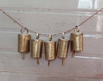 Puede incluir: Una cadena de seis campanas doradas colgando de una cuerda de colores. Las campanas son cilíndricas y tienen una parte superior ligeramente redondeada.