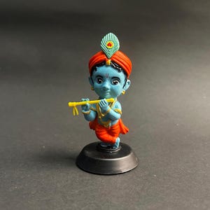 Könnte beinhalten: Figur einer blauhäutigen Gottheit, geschmückt mit einem roten Turban und orangefarbener Kleidung, die eine gelbe Flöte hält. Eine Pfauenfeder ziert den Kopf. Die Figur steht auf einem schwarzen Sockel vor dunklem Hintergrund.