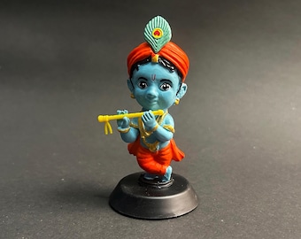 May include: A blue-skinned deity figurine, wearing a red turban and orange garments, holding a yellow flute. A peacock feather decorates the head. The figurine is on a black base, against a dark backdrop.