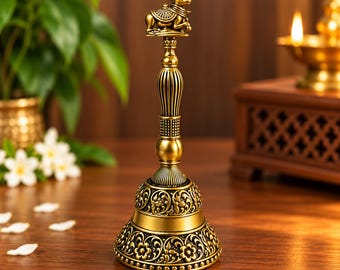 May include: A metallic gold-toned hand bell featuring an intricate floral pattern embossed on the base and a sculpted figure of a reclining bull at the top. The handle has a ribbed texture and a tapered design, leading to a wide, flared bell body.